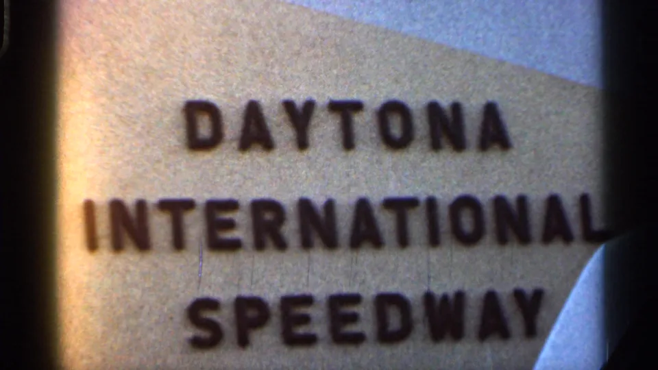 Close 8mm view of the Daytona International Speedway sign, with a vehicle edge and gate shadow framing the shot.