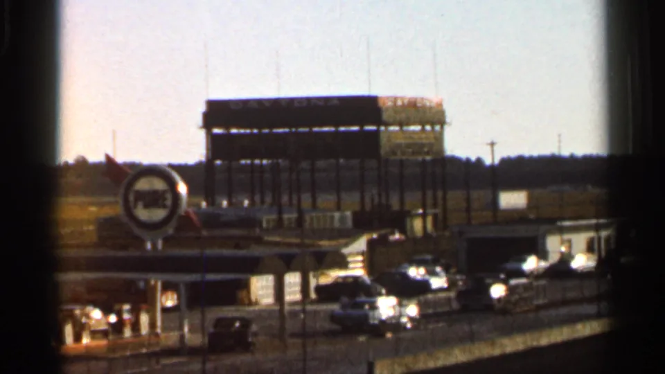 Wide 8mm frame of the Daytona area with Pure Oil station signage, classic cars, and a grandstand in the background.
