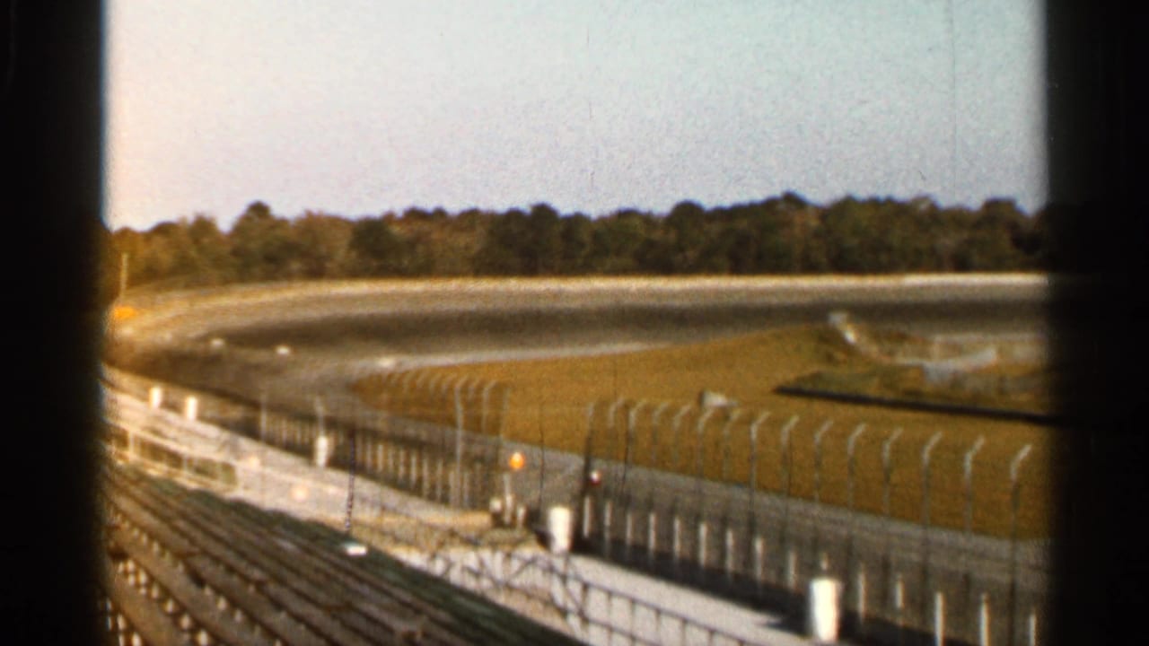 1966 Florida home-movie frame showing a curving racetrack, grass verge, safety fence, empty seating, and a distant tree line.