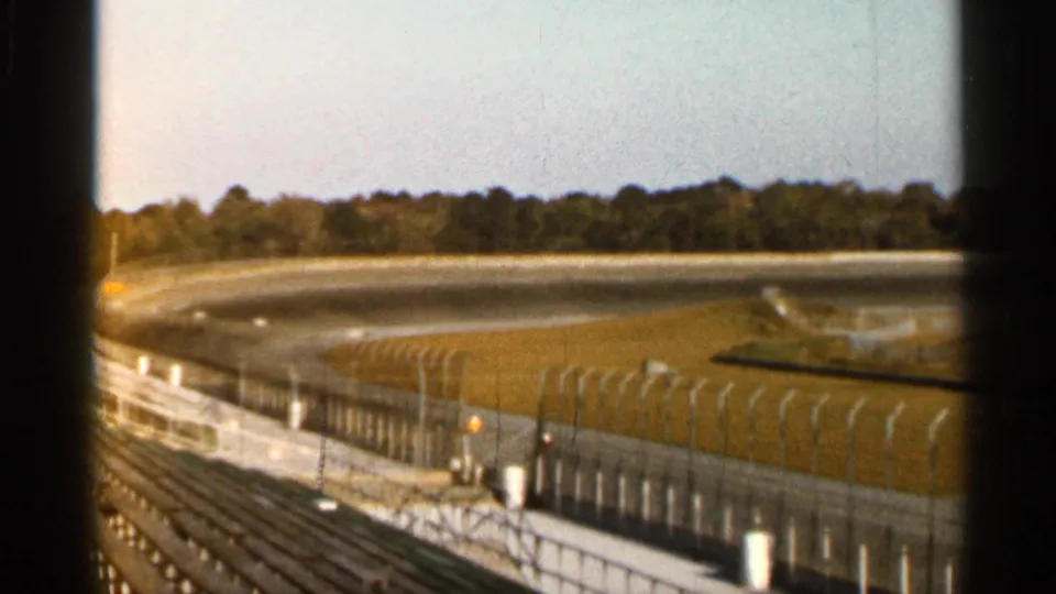 1966 Florida home-movie frame showing a curving racetrack, grass verge, safety fence, empty seating, and a distant tree line.