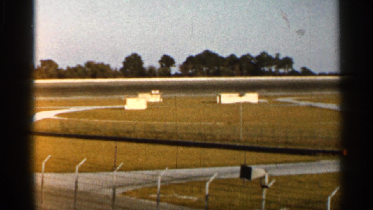 1966 Florida 8mm frame of a racetrack infield with white boxes or signs, low fencing, track surface, and trees beyond.
