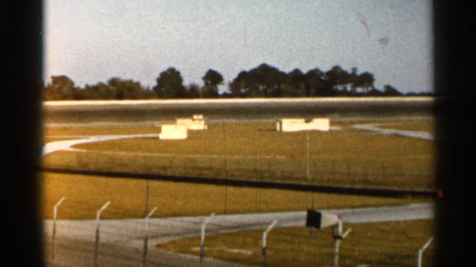 1966 Florida 8mm frame of a racetrack infield with white boxes or signs, low fencing, track surface, and trees beyond.