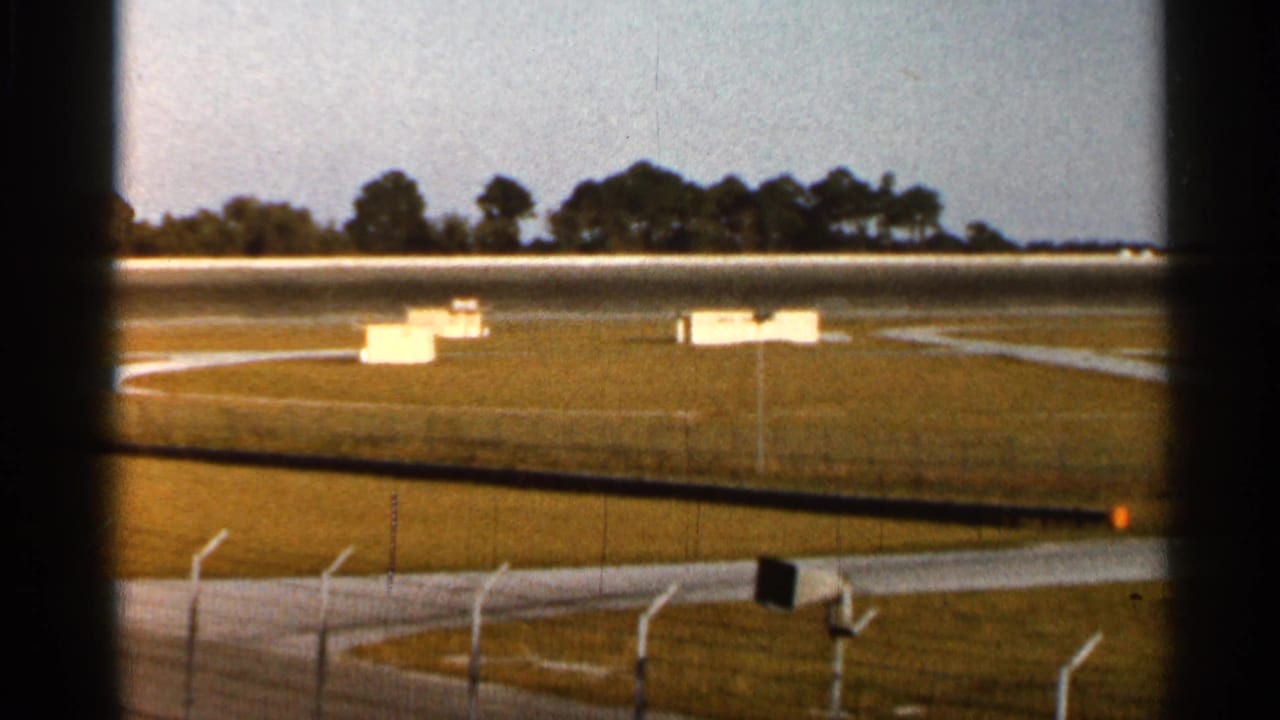 1966 Florida home-movie view across a racetrack fence toward grass infield, white signs or boxes, asphalt lanes, and distant trees.