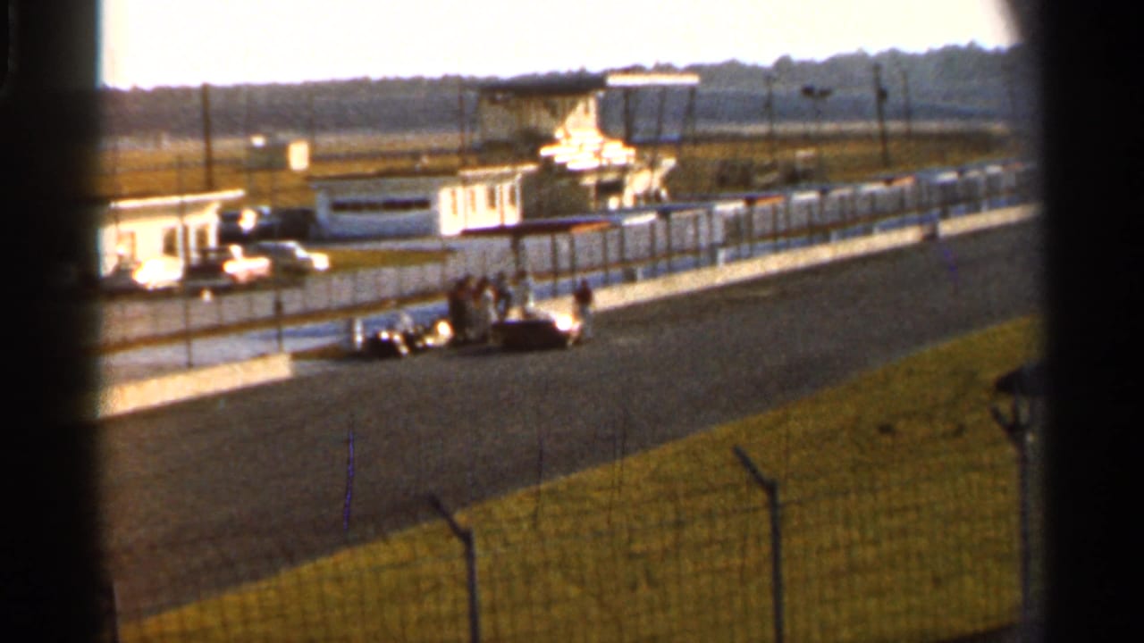 1966 Florida racetrack scene with a vintage race car near the pit wall, several people nearby, and grandstand or control structures behind.