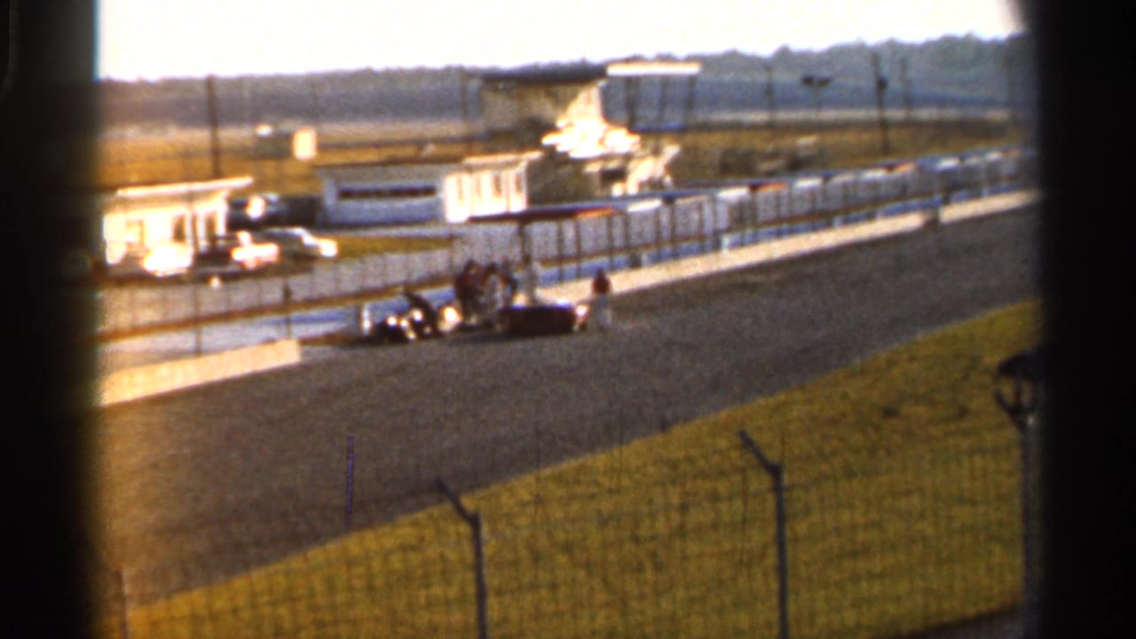 Handheld 8mm view of a vintage race car at a Florida racetrack, with people standing nearby and pit-side buildings in the background.