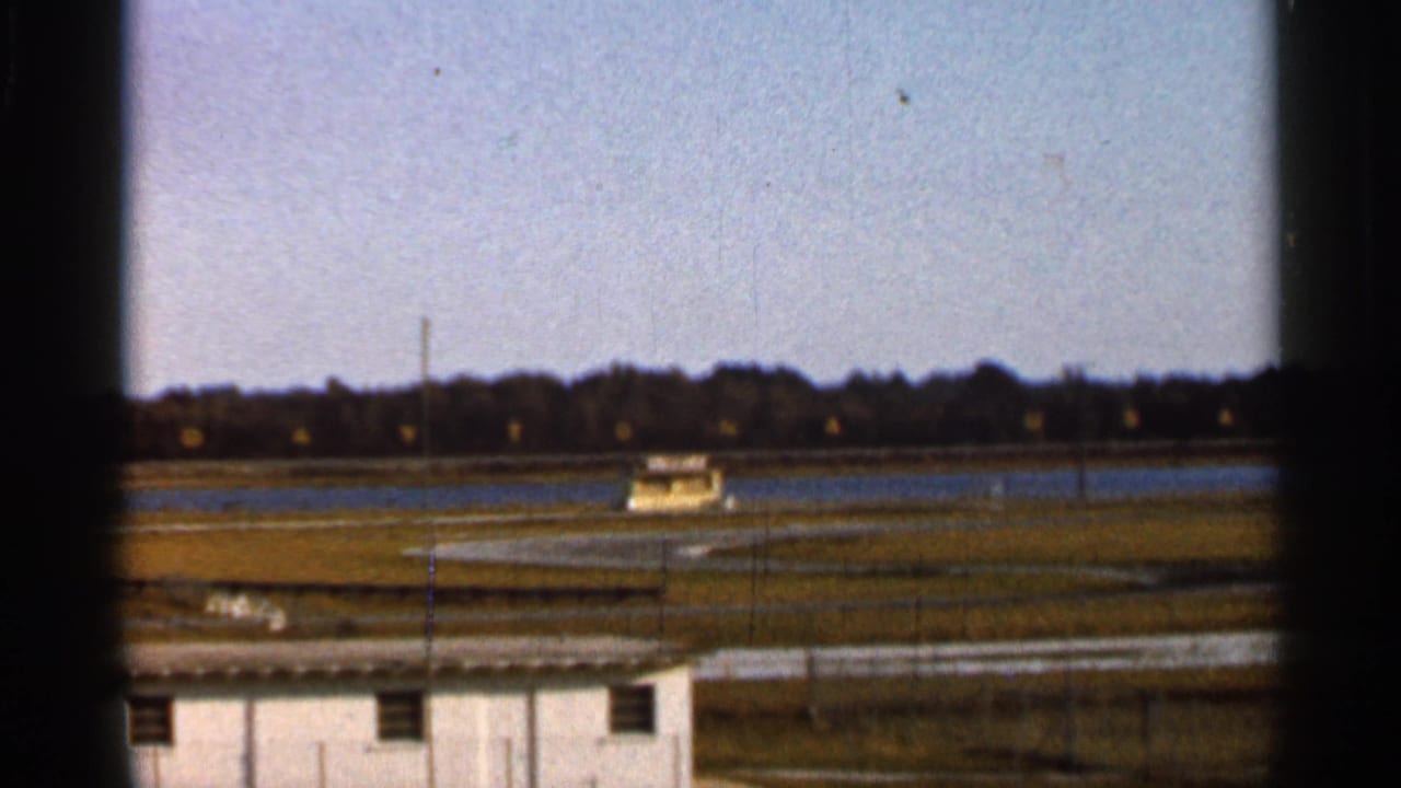 1966 Florida racetrack view showing a distant water feature, small building, track lanes, grassy areas, and treeline under a bright sky.
