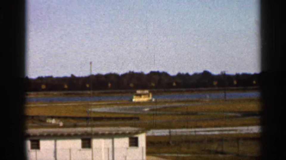 1966 Florida 8mm frame showing a distant race car on a track, viewed past a fence with a small white building in front.
