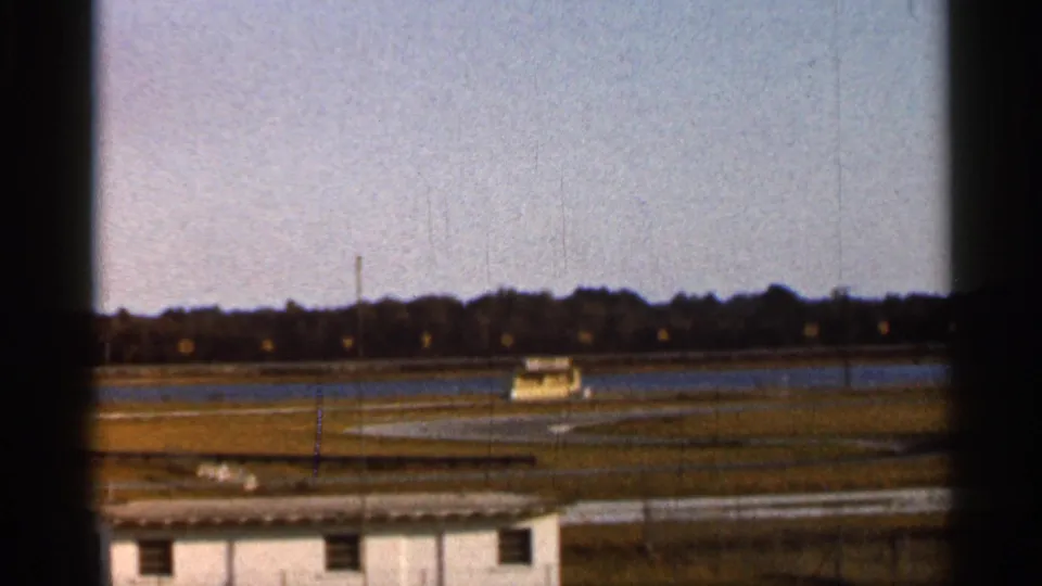 1966 Florida racetrack view with a small race car in the distance, framed by fence posts, grass, and trees.
