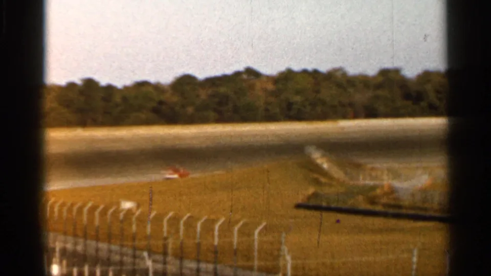1966 Florida 8mm frame with a red race car and a white car on a track beyond a curved safety fence.