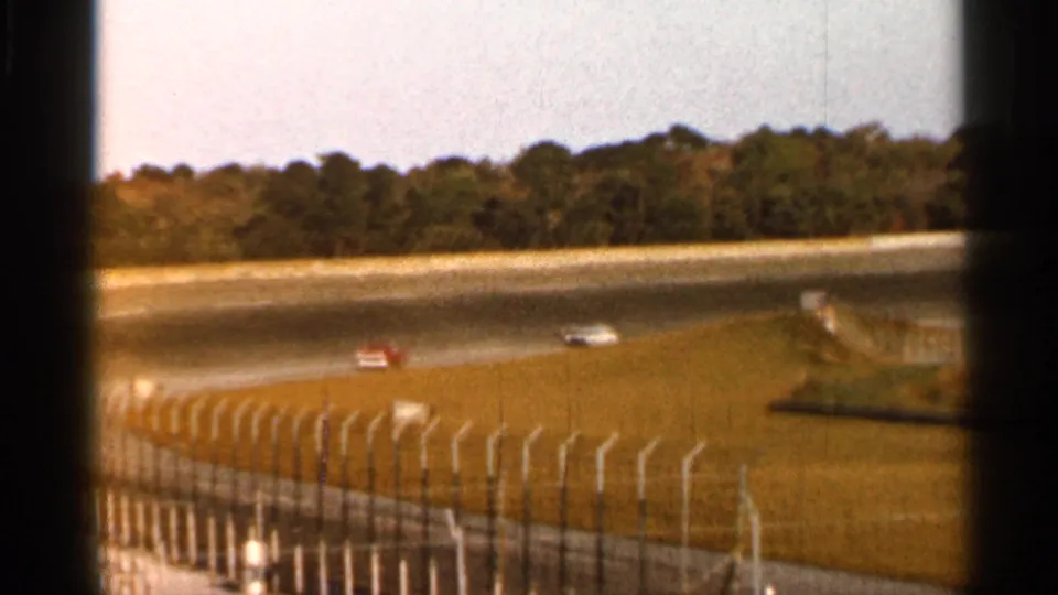 1966 Florida racetrack scene showing red and white cars on the far track, seen over grass and safety fencing.