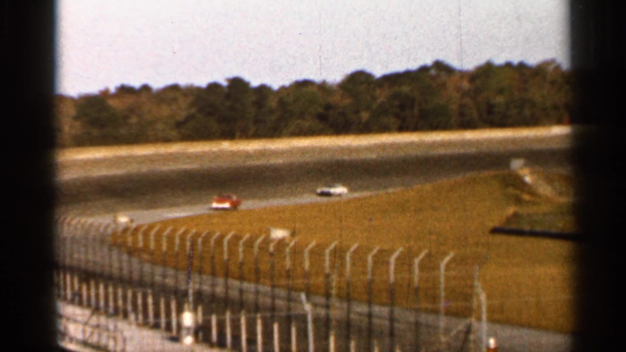 1966 Florida 8mm view of race cars on a curved track, seen through dark foreground framing with fencing, grass, and trees beyond.