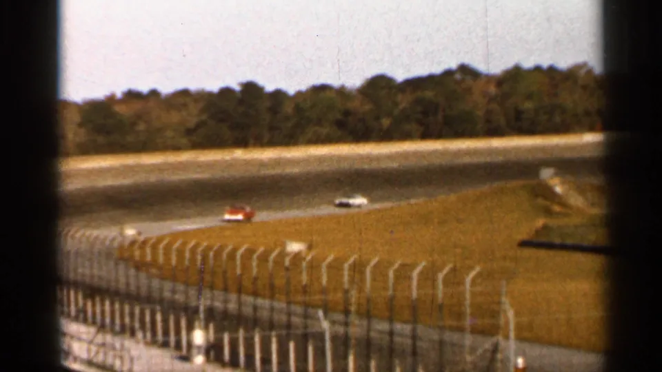 1966 Florida 8mm view of race cars on a curved track, seen through dark foreground framing with fencing, grass, and trees beyond.