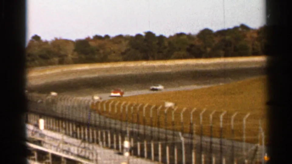 1966 Florida home-movie frame showing cars spaced along a circular race track, with a fence, infield grass, and tree line visible.