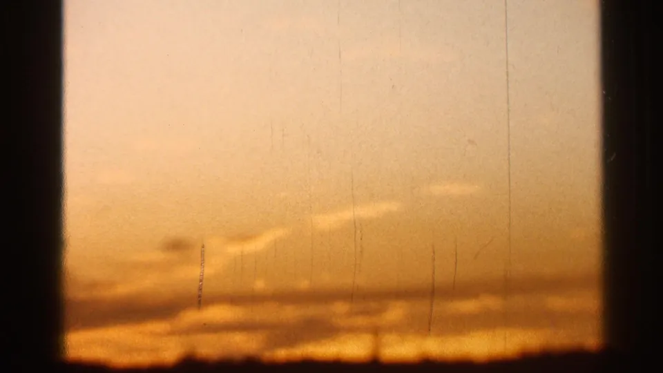 Warm orange sunset sky on 8mm film, with soft cloud bands, vertical film marks, and a dark low horizon.