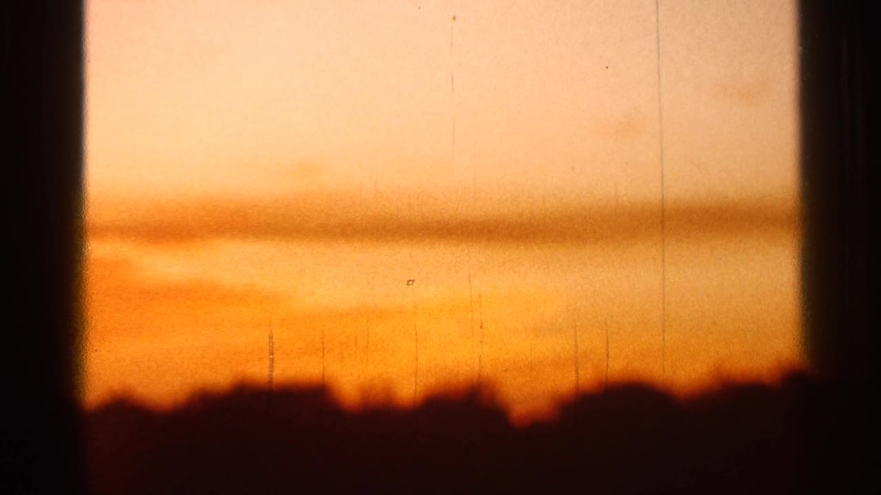 1966 Florida 8mm frame showing a warm orange dawn sky, dark low horizon, soft haze, grain, scratches, and dark frame-edge shadows.