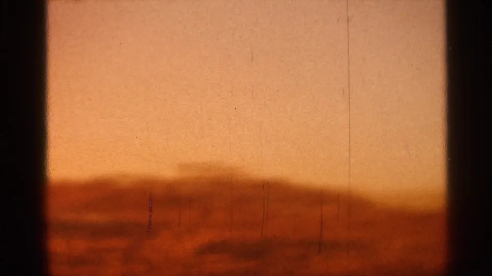1966 Florida 8mm frame with amber dawn light over a blurred low horizon, visible grain, vertical scratches, and heavy dark edge shadows.