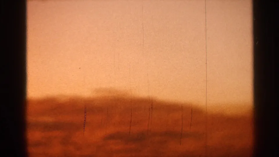1966 Florida 8mm frame showing pale orange dawn sky, reddish low horizon, faint haze, vertical scratches, grain, and dark gate shadows.