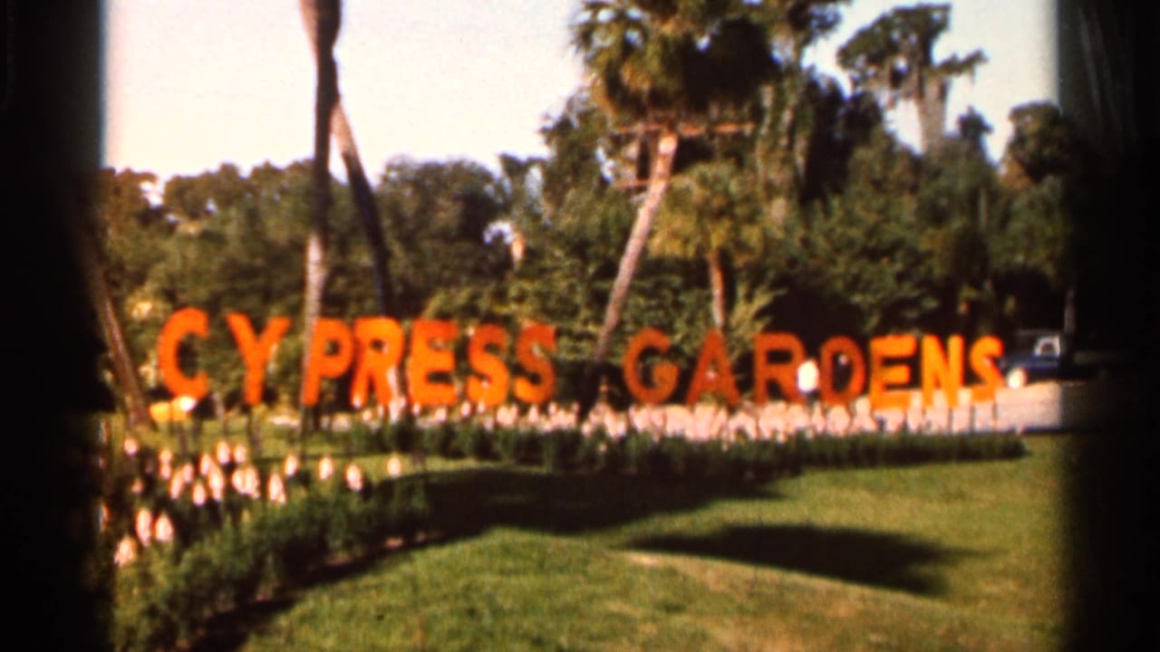 Orange Cypress Gardens sign across a lawn with flower beds, palms, and trees behind it.