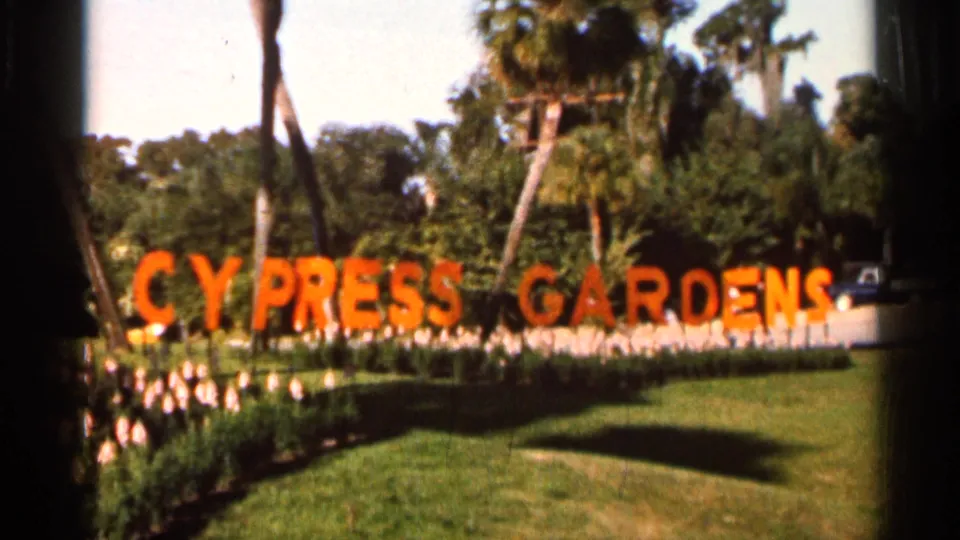 1966 Florida home-movie view of the Cypress Gardens sign with landscaped lawn, palms, flowers, and a car partly visible beyond the sign.