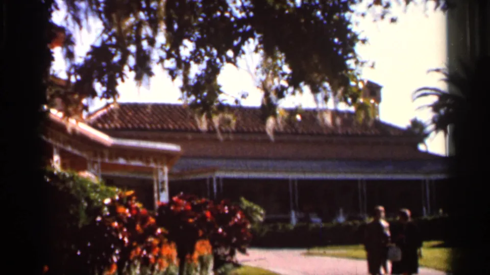 1966 Florida 8mm frame showing a stately tiled-roof building at Cypress Gardens, with flowering shrubs, mossy trees, and two women visitors.