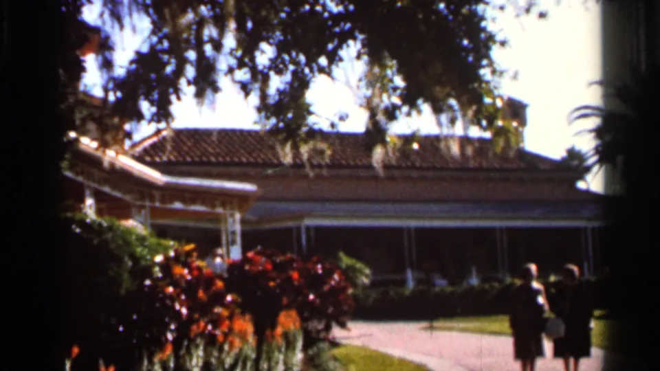 1966 Florida home-movie frame of the Cypress Gardens building and porch, framed by Spanish moss, palms, red shrubs, and women visitors on the path.