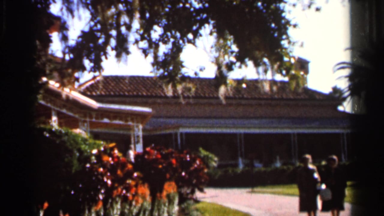 Tiled-roof house seen through trees, with two figures near a driveway.