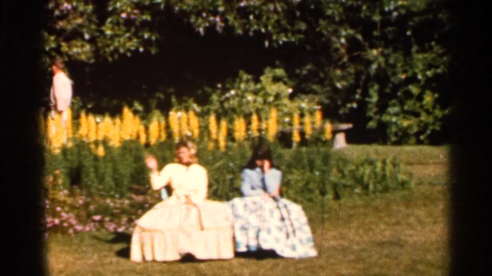 Two women seated outdoors on a Florida lawn in 1966, with one waving near tall yellow flowers and shrubs.