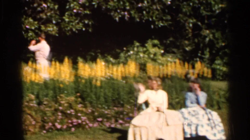 Garden scene from 1966 Florida home-movie footage with two seated women and another adult standing near tall yellow flowers.