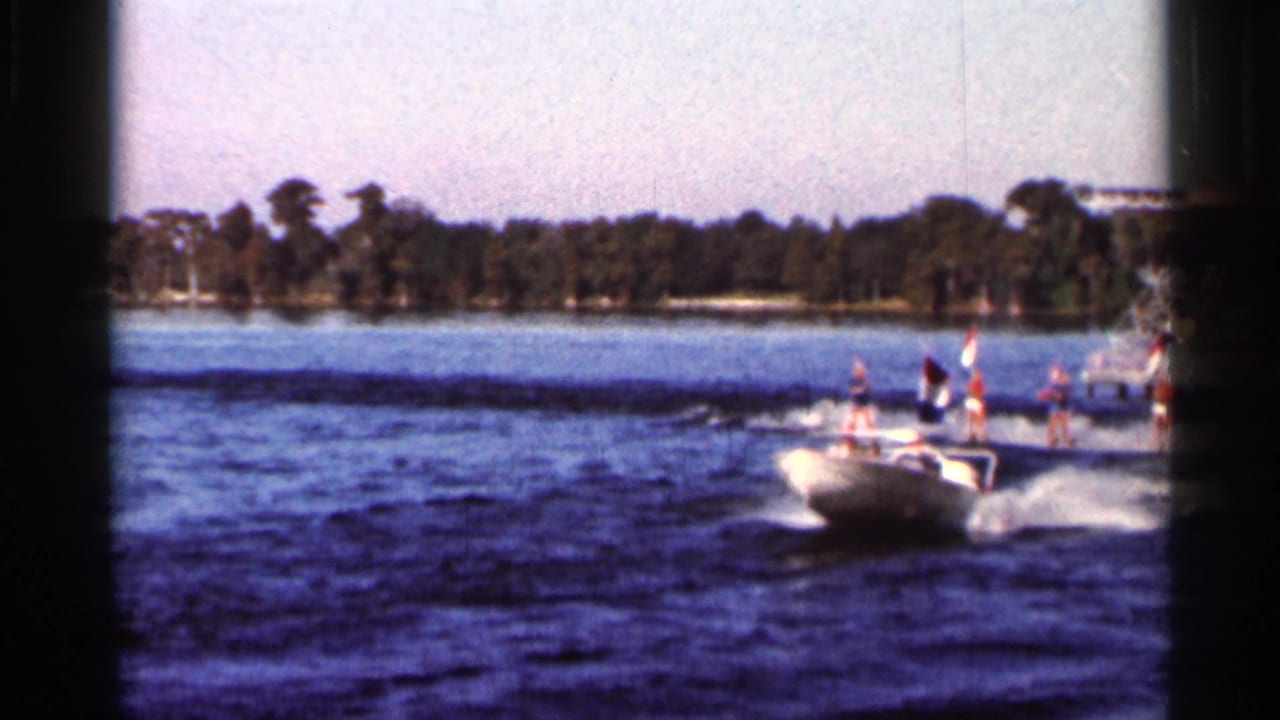 Speedboat pulls flag-carrying water skiers across a lake near a wooded shoreline.