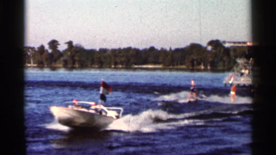 The boat continues across the Florida lake as distant skiers follow in its wake, each appearing to hold a small flag.