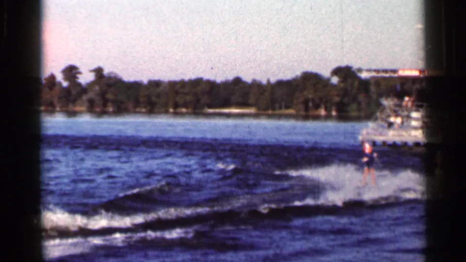 A distant water skier moves through spray near a larger boat, with the wooded Florida shoreline behind.