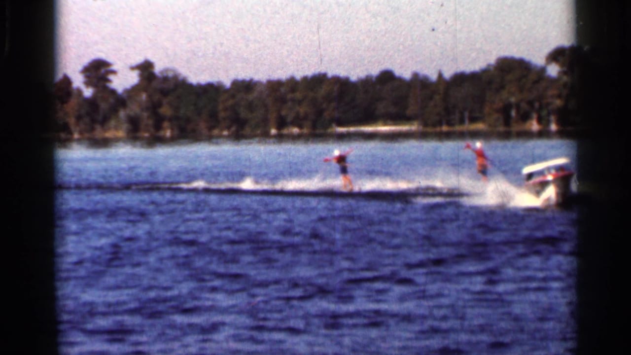 1966 Florida 8mm frame showing two distant water skiers being pulled by a small motorboat across open water near a wooded shoreline.