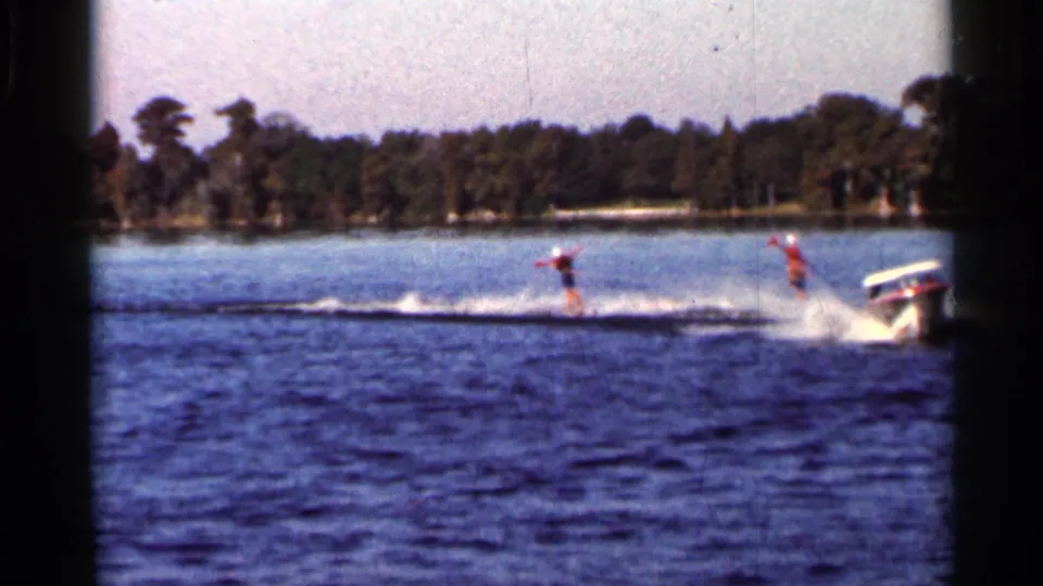 1966 Florida 8mm frame showing two distant water skiers being pulled by a small motorboat across open water near a wooded shoreline.