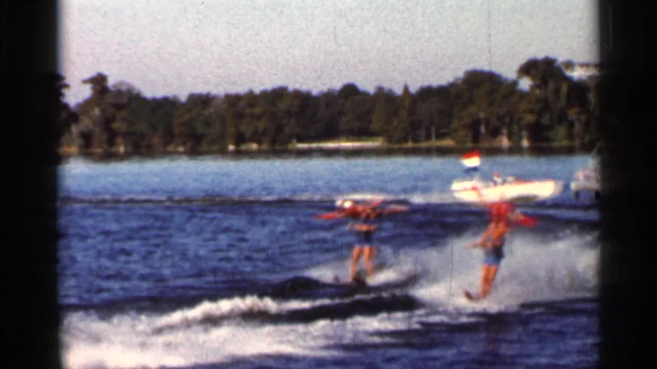 1966 Florida home-movie frame with water skiers crossing the foreground and a small motorboat nearby on a sunny lake or inlet.