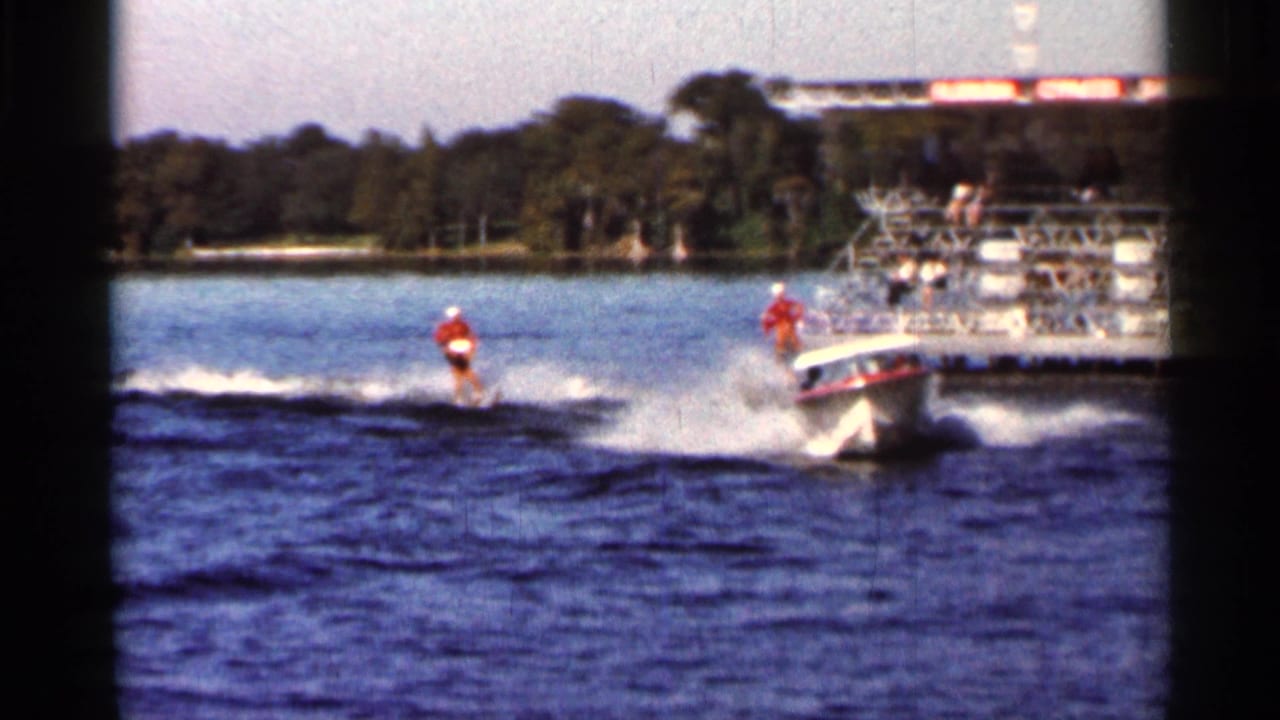 Two water skiers trail a speedboat on a lake near a dock and trees.