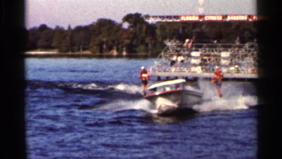 Water-ski performers follow a turning speedboat on a Florida lake, with Cypress Gardens signage in the background.