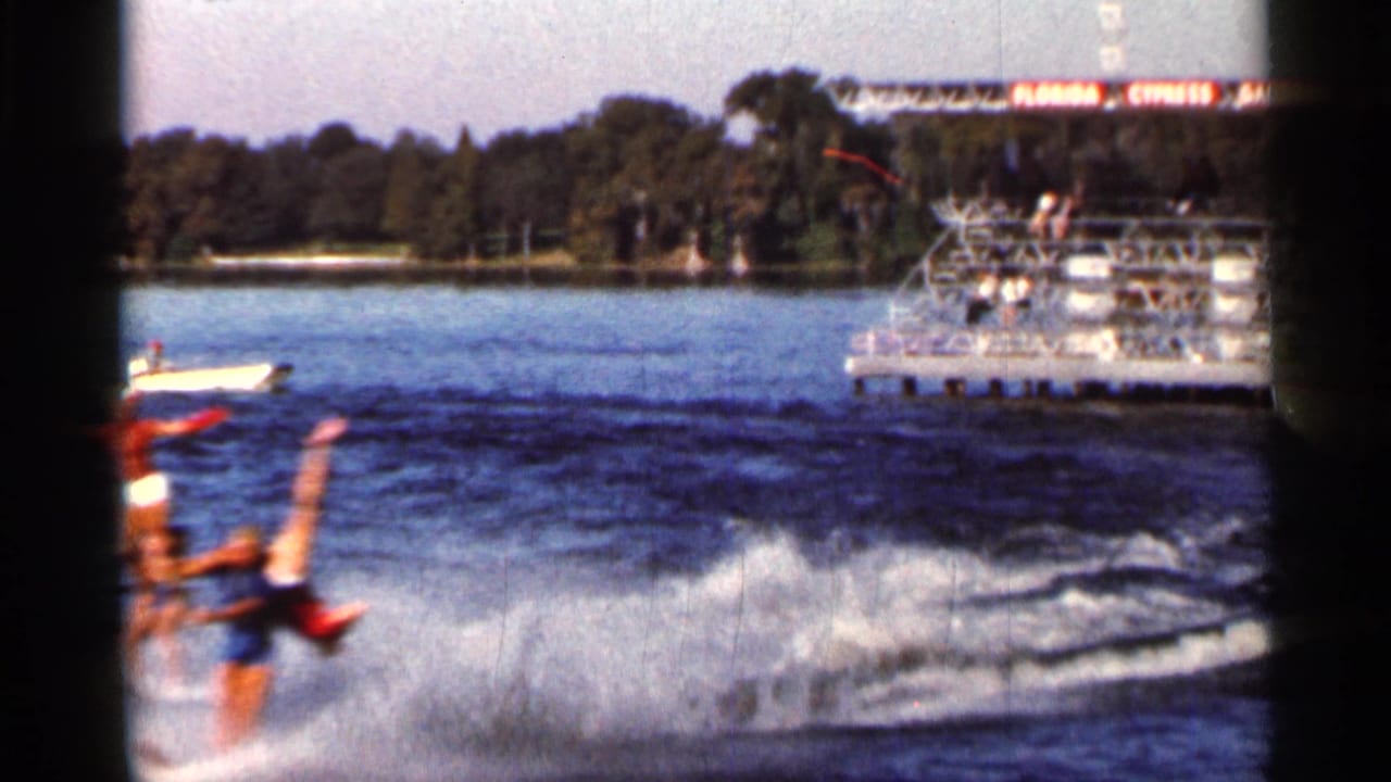 Two water skiers pass across a lake with a spectator pier and sign in the background.