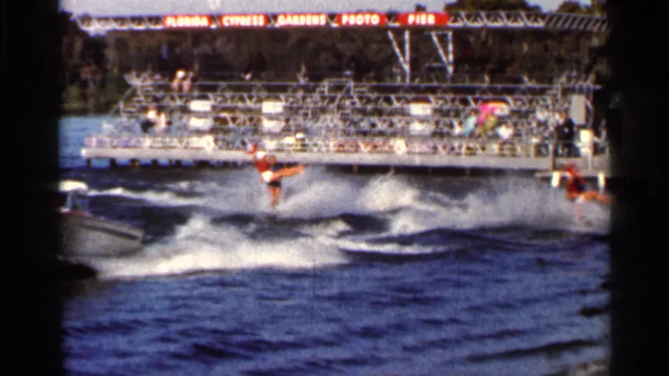 Synchronized skiers continue across the lake near the Cypress Gardens photo pier, with spectators visible behind them.