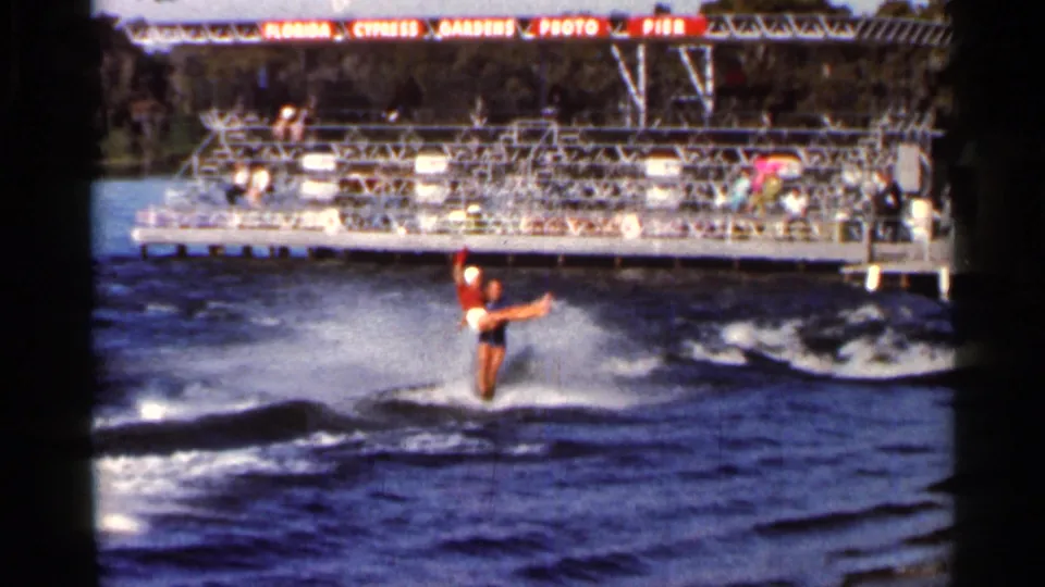 A performer in red swimwear skis with arms extended on a Florida lake near the Cypress Gardens spectator pier.