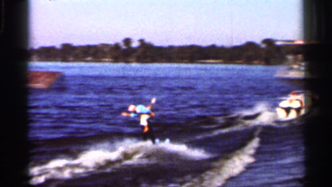 Water skier in a blue shirt rides behind a speedboat on a lake.