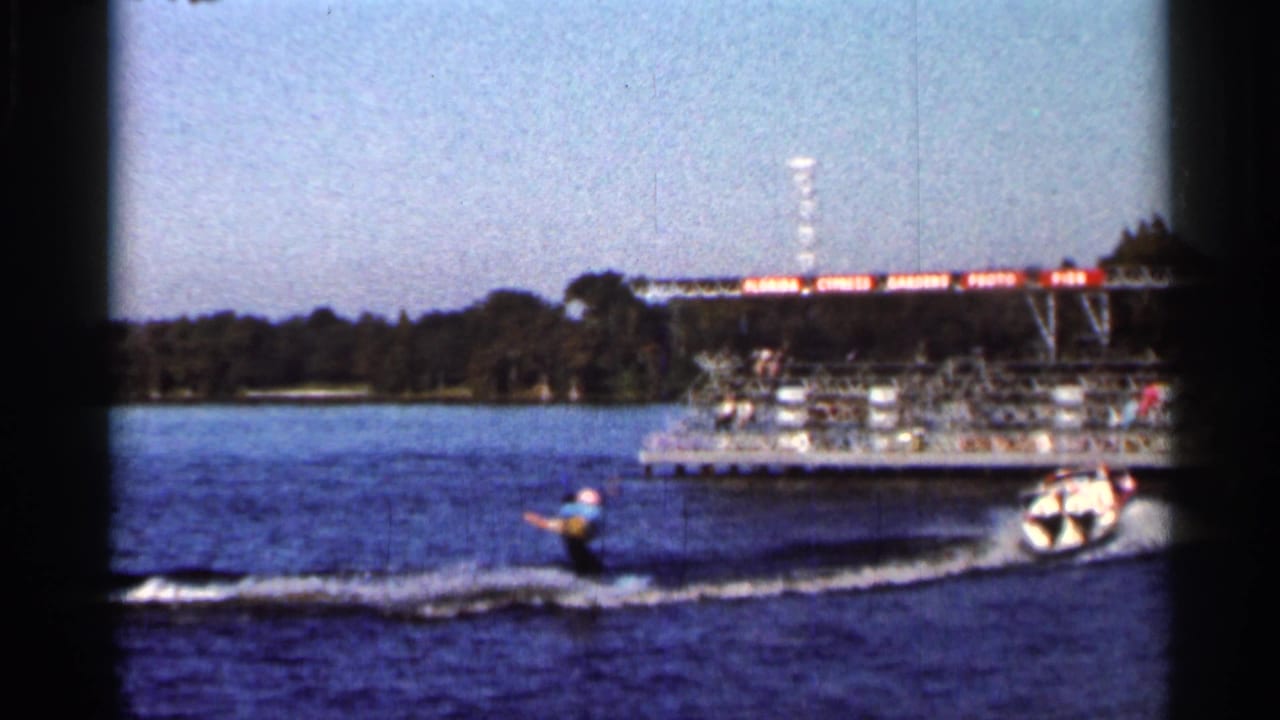 Water skier trails a small speedboat past a dock area in 1966 Florida lake footage.