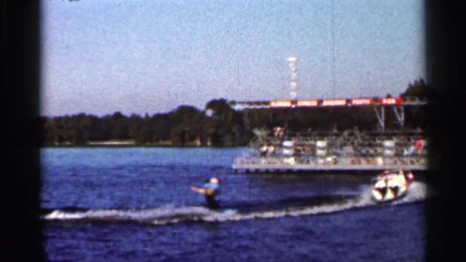 Water skier trails a small speedboat past a dock area in 1966 Florida lake footage.
