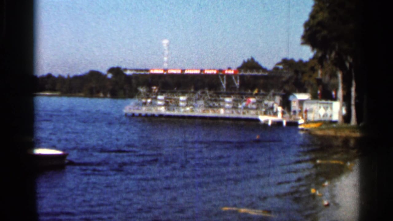 Wide 1966 Florida lakeside view with a docked platform, trees along the shore, and tow wake crossing the water.