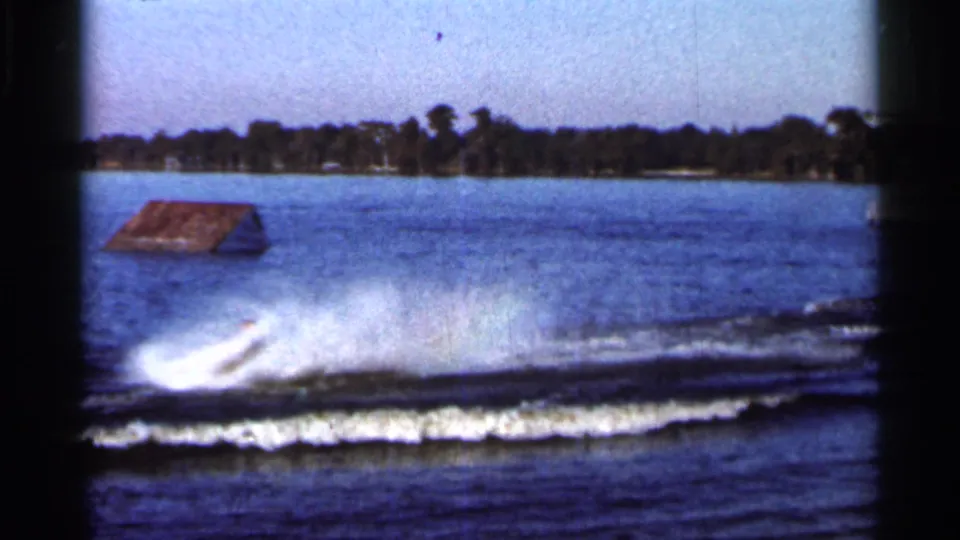 1966 Florida lake footage with white waterski spray, dark wake lines, and a low ramp-like object near the far shoreline.