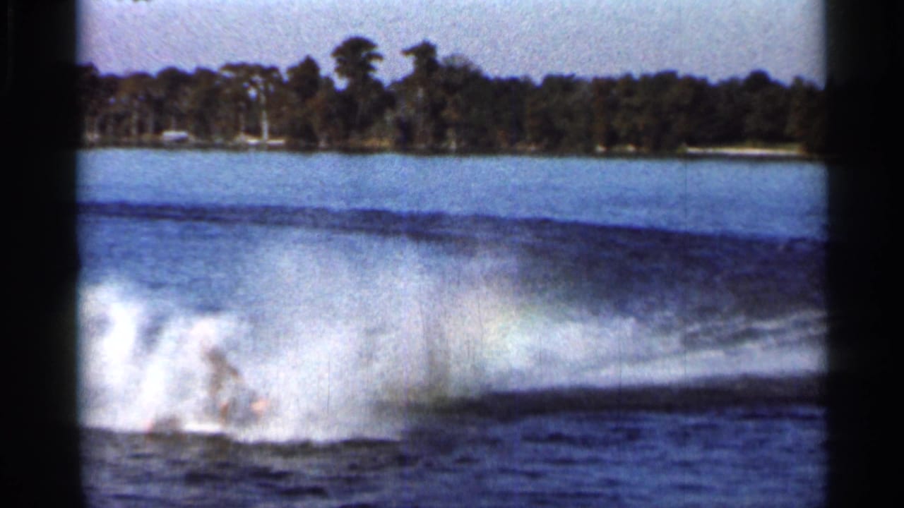 Water skier partly obscured by spray on a lake with a tree-lined shoreline behind.
