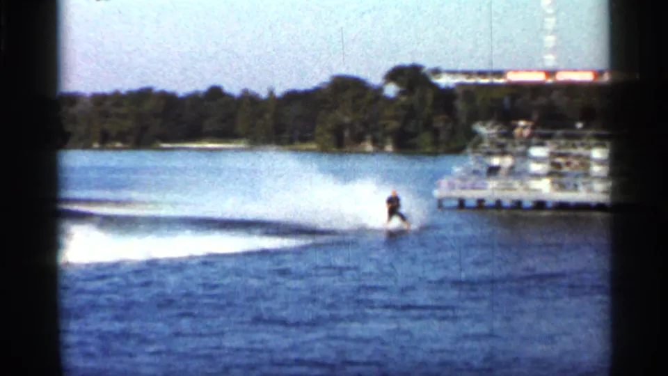 The skier moves across open water with a long wake, with Cypress Gardens dock structures and trees in the background.
