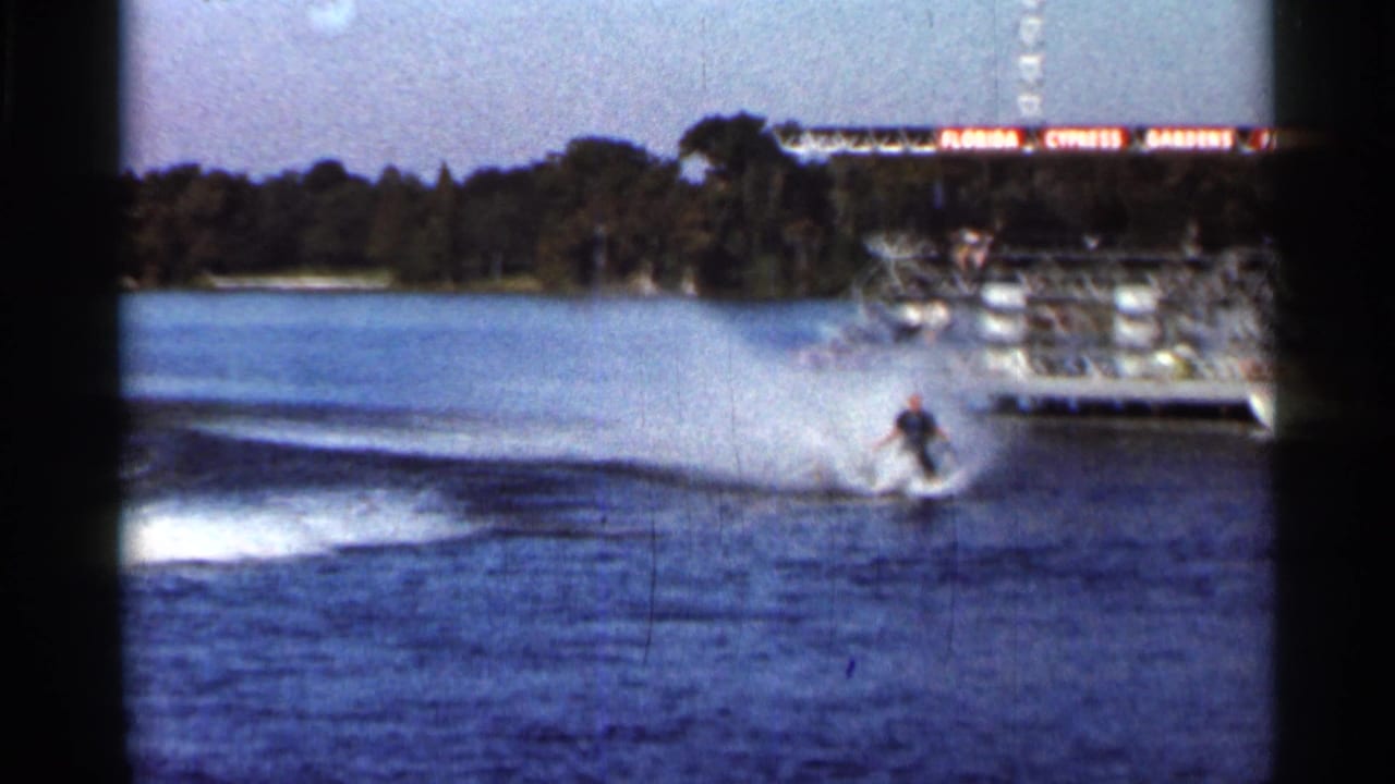 1966 Florida 8mm frame showing a water skier moving across a lake near the Florida Cypress Gardens Photo Pier, with spray, shoreline trees, and film gate borders visible.