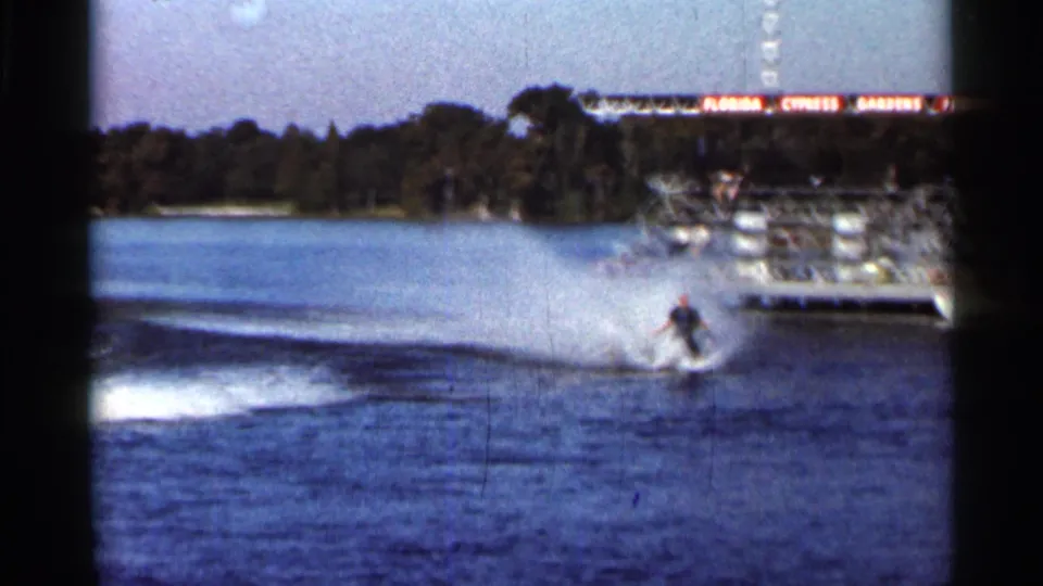 1966 Florida 8mm frame showing a water skier moving across a lake near the Florida Cypress Gardens Photo Pier, with spray, shoreline trees, and film gate borders visible.