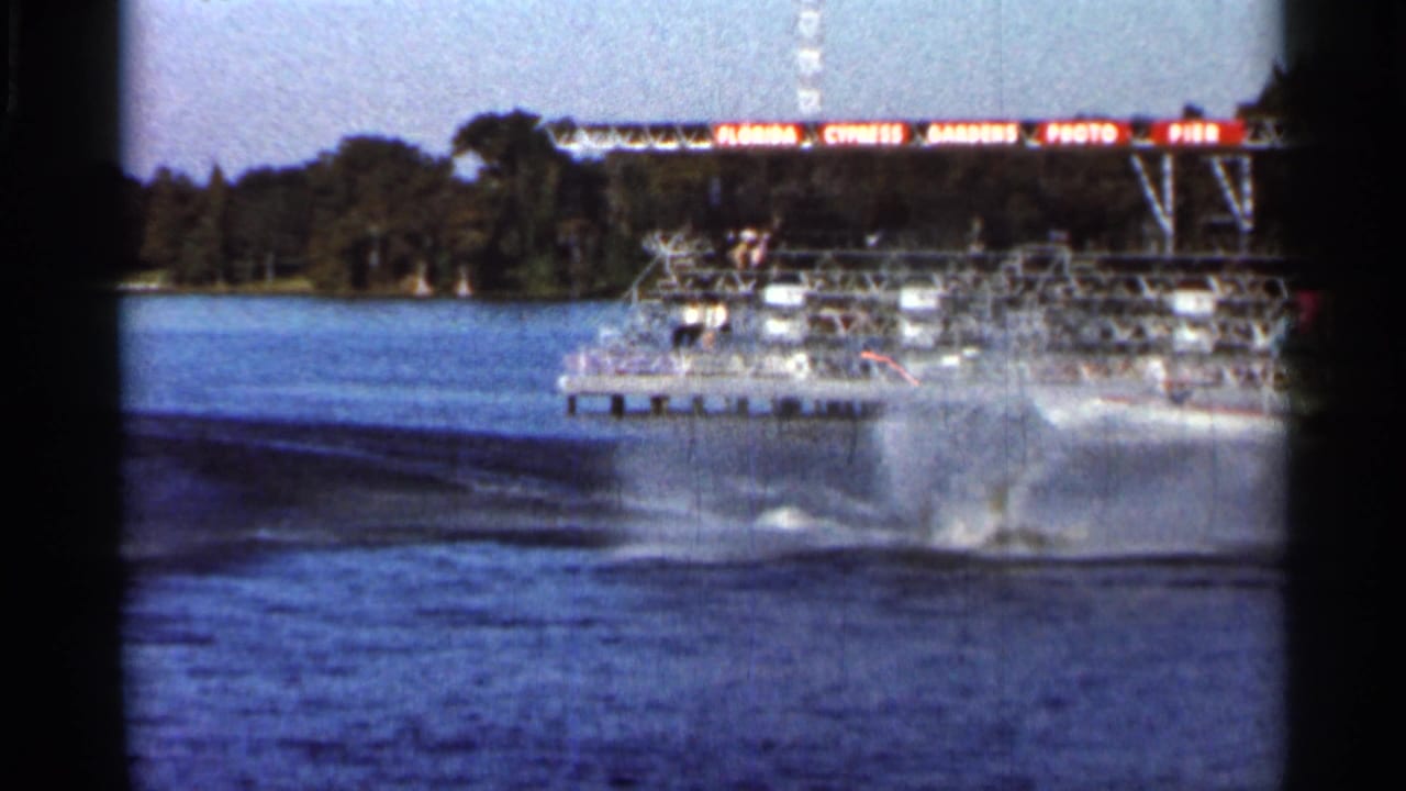 Speedboat wake and spray cross the lake near a viewing pier and shoreline trees.