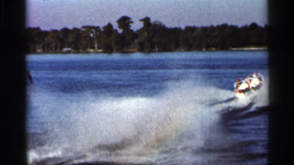 1966 Florida 8mm frame showing a speedboat cutting across the lake with heavy white spray, blue water, wooded shoreline, and dark home-movie frame edges.